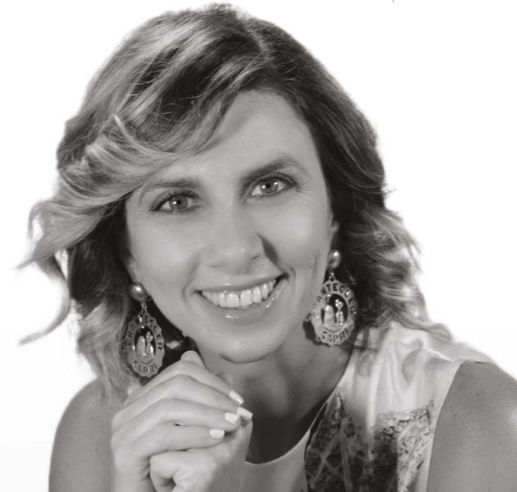 Una donna con capelli ondulati e lunghi fino alle spalle sorride alla telecamera, indossando grandi orecchini rotondi e un top senza maniche a fantasia. Appoggia il mento sulla mano su uno sfondo semplice. La foto è in bianco e nero.