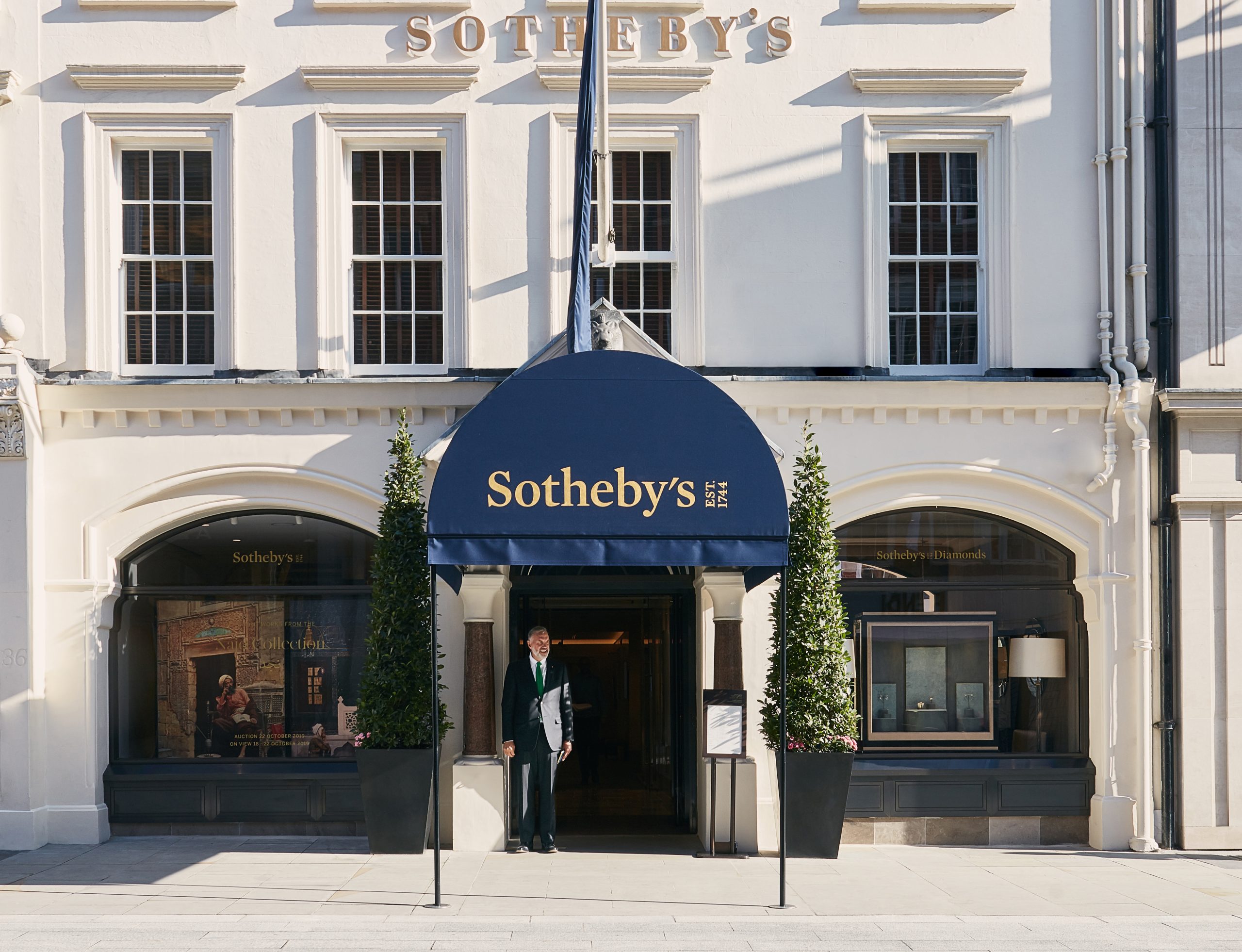 La facciata di un edificio di Sotheby's con una tenda blu sopra l'ingresso allude alle vivaci vendite 2024. Grandi finestre fiancheggiano la porta e un portiere in giacca e cravatta è in piedi sotto la tenda. Due alte fioriere con verde sono posizionate vicino all'ingresso.