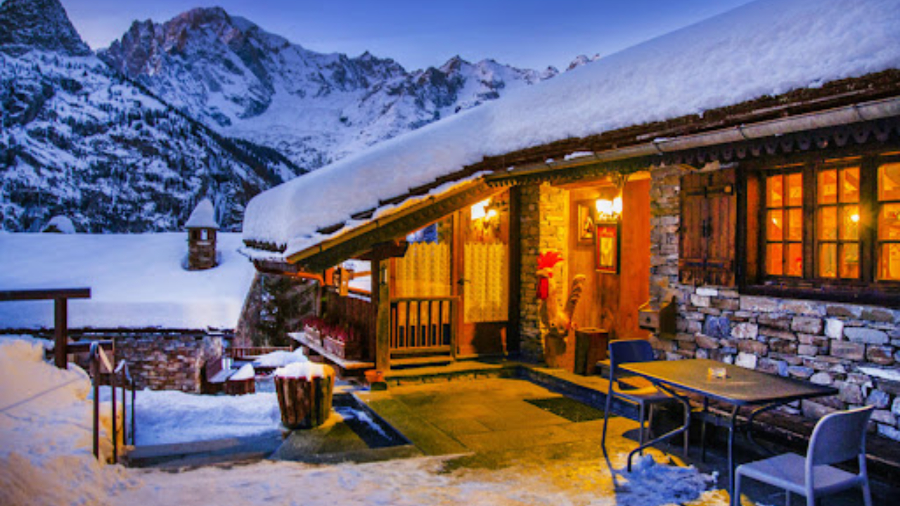 Accogliente baita di montagna con luci calde che brillano all'interno, circondata da un paesaggio innevato. Struttura rustica in legno con un piccolo portico e posti a sedere all'aperto, sullo sfondo di maestose cime innevate al crepuscolo.