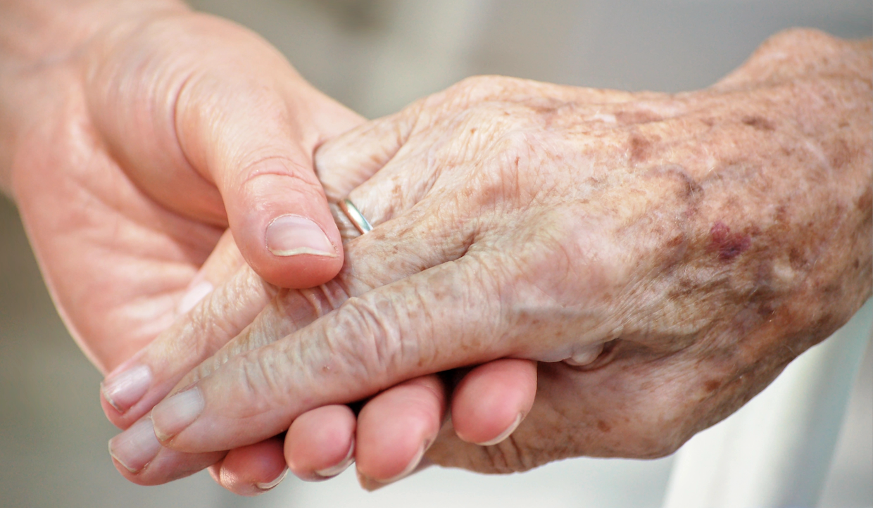 Un primo piano di una giovane mano che tiene e sostiene delicatamente una mano anziana con macchie di età visibili e una fede nuziale. L'immagine trasmette temi di cura, supporto e connessione intergenerazionale.