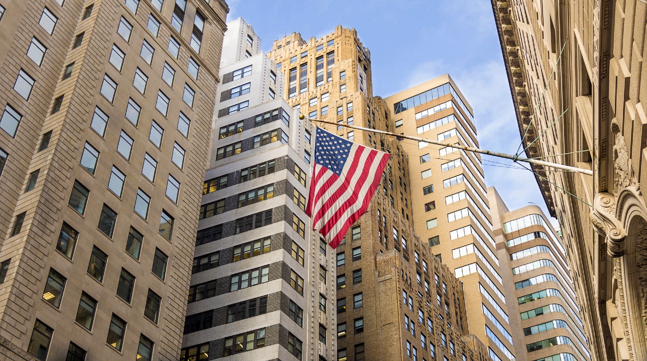 Una grande bandiera americana è appesa tra alti grattacieli in una città. Gli edifici hanno vari stili architettonici, con facciate in vetro e pietra. Il cielo è limpido e azzurro, e proietta luce naturale sulla scena urbana.