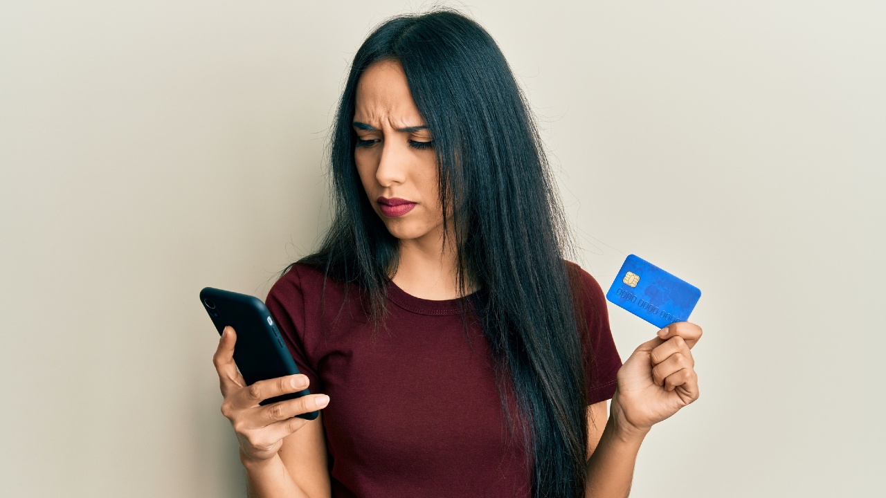 Una donna con lunghi capelli scuri e una camicia rossa sembra preoccupata, tiene in mano uno smartphone e una carta di credito blu e contempla un bonifico istantaneo su uno sfondo semplice.