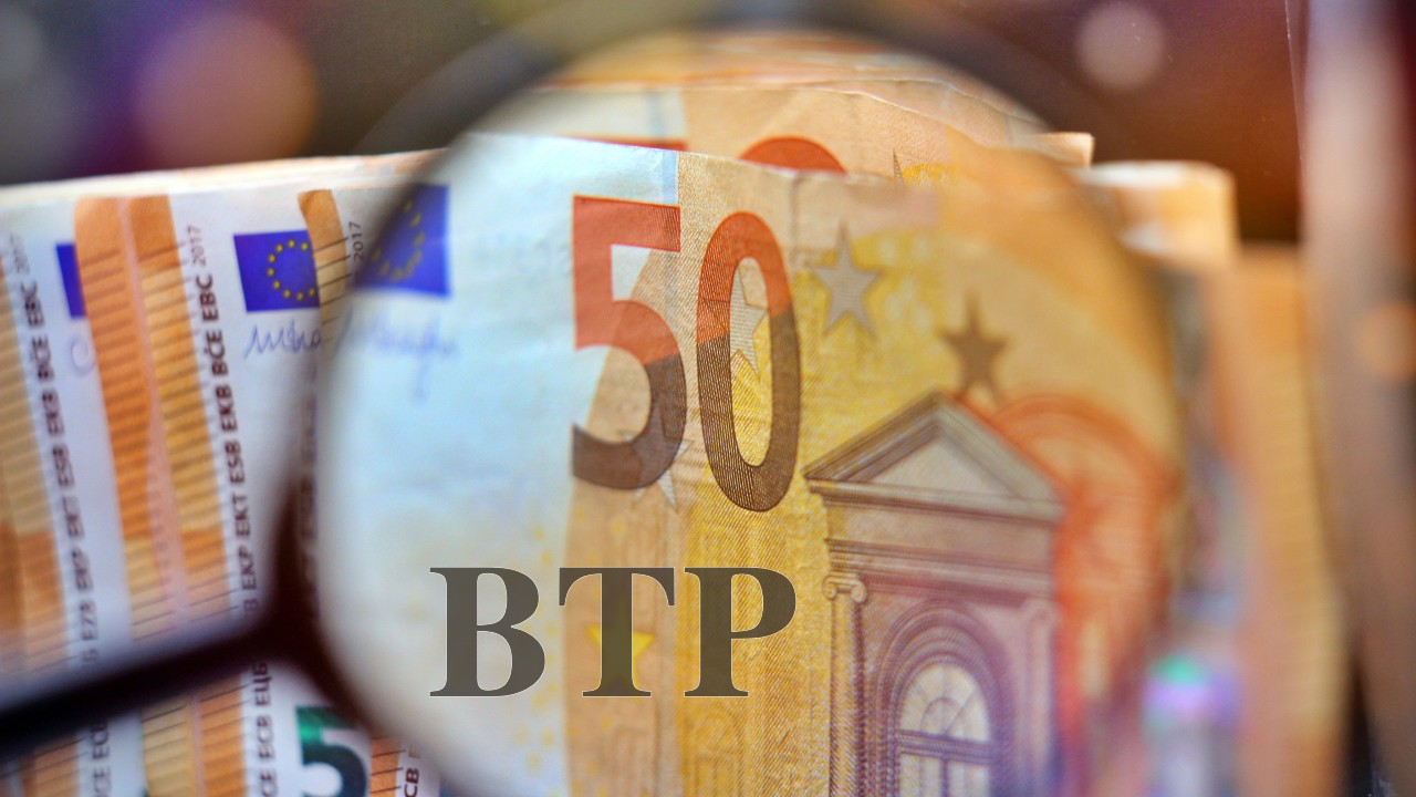 Una lente di ingrandimento si concentra su una pila di banconote da 50 euro, con le lettere "BTP" sovrapposte all'immagine. Lo sfondo rivela un primo piano degli intricati disegni architettonici delle banconote.