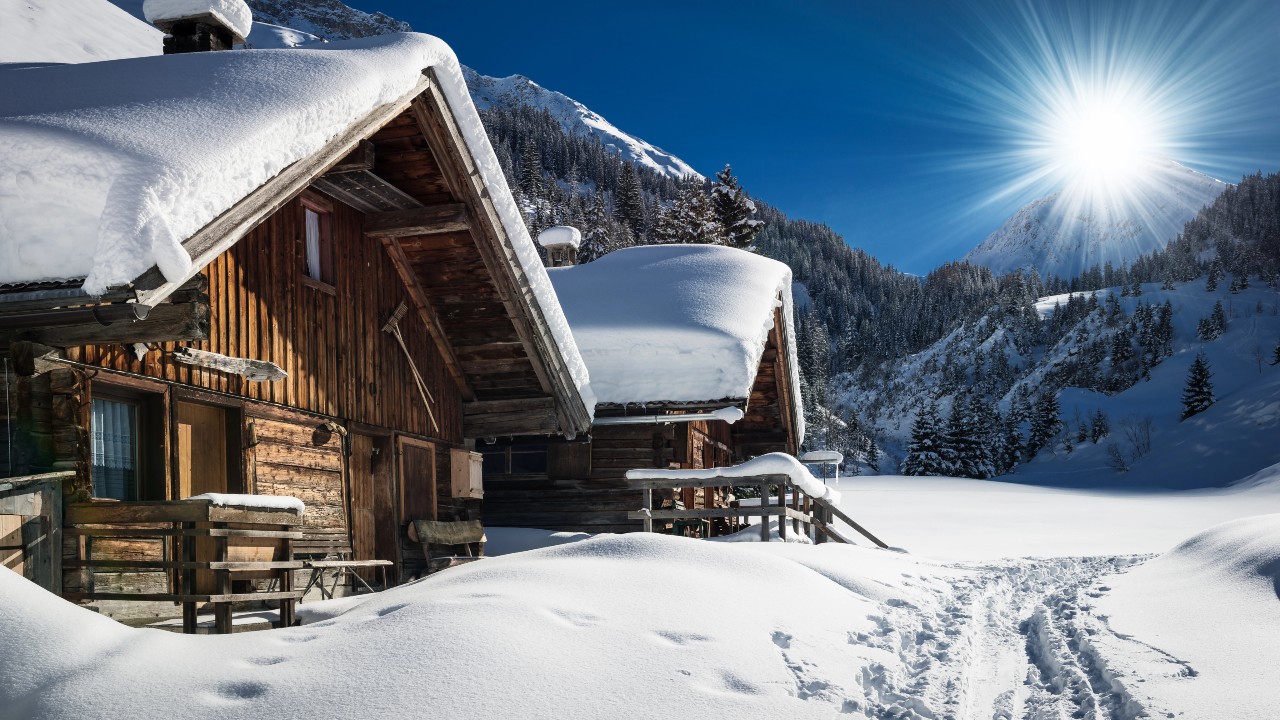 Una casa in montagna coperta di neve si trova in uno spettacolare paesaggio invernale, circondata da pini e vette sotto un cielo azzurro e terso. Il sole splende luminoso, proiettando lunghe ombre sulla neve intatta. Un sentiero conduce verso questi invitanti rifugi.
