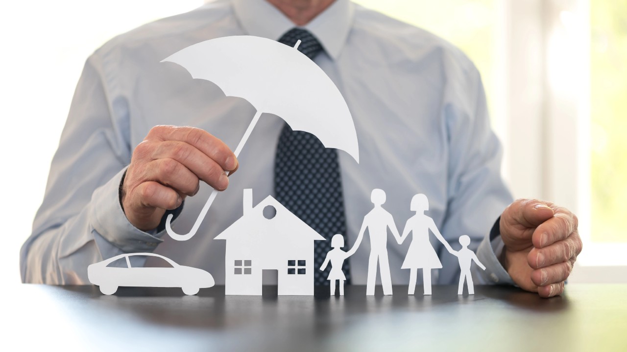 Una persona in giacca e cravatta tiene in mano una grande figura con un ombrello sopra ritagli di carta di un'auto, una casa e una famiglia, a simboleggiare la protezione e la sicurezza per i beni personali e le persone care. Questa immagine è usata per chiedersi se sia meglio una donazione o un patto di famiglia per la protezione del patrimonio.