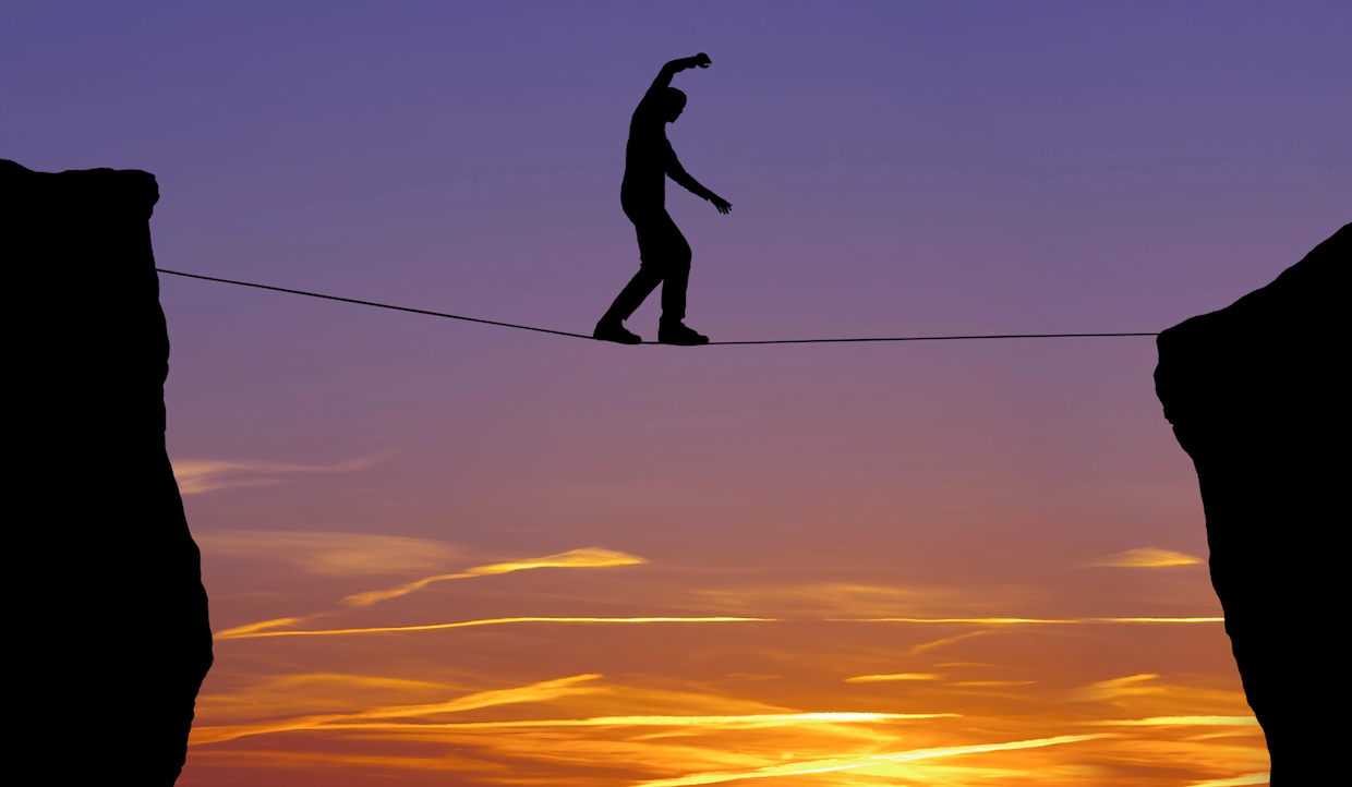 La sagoma di una persona in equilibrio su una fune tra due scogliere al tramonto, con un cielo dai colori vivaci, arancioni e viola sullo sfondo.