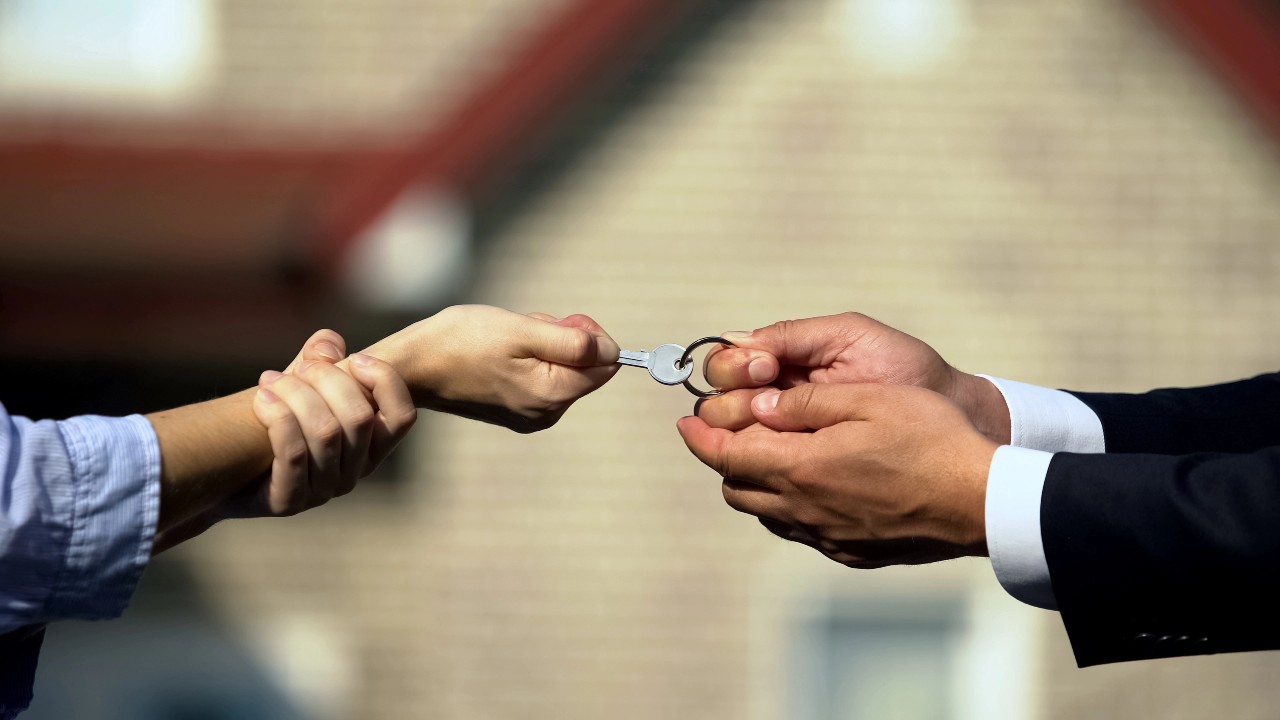 Primo piano di due mani che si scambiano una chiave. Una mano, che indossa un abito, porge la chiave con un portachiavi a forma di casa verso un'altra mano con una camicia casual, suggerendo una transazione immobiliare o di affitto. Un edificio sfocato è sullo sfondo. L'immagine è usata per far riferimento a un trasferimento di eredità.