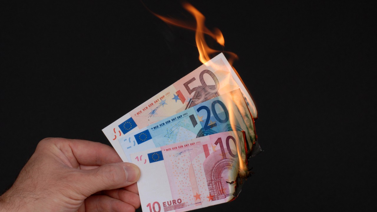 Una mano che tiene delle banconote in euro in fiamme, tra cui quelle da 50, 20 e 10 euro, su uno sfondo nero. Le fiamme consumano visibilmente i bordi delle banconote, una metafora della perdita finanziaria e della perdita dell’esenzione fiscale.