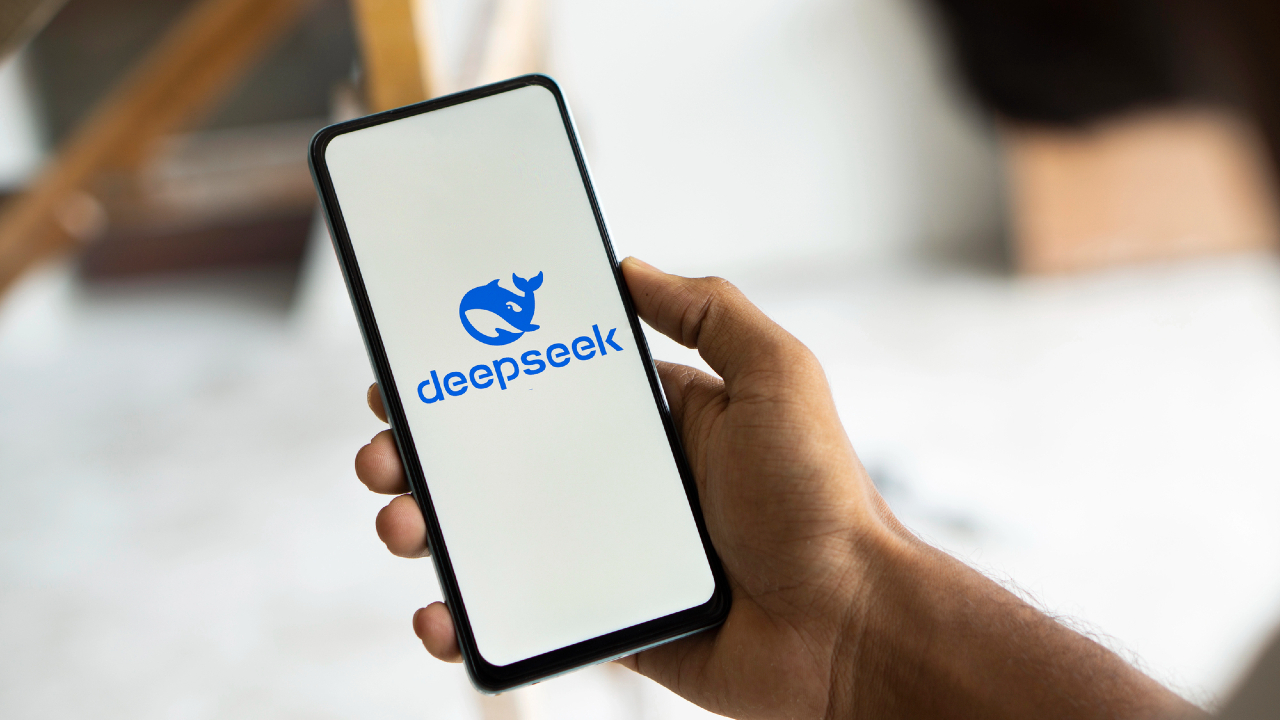 Una mano afferra uno smartphone, che mostra in modo evidente il logo "deepseek" raffigurante un rinoceronte blu su uno schermo bianco nitido. Lo sfondo sfocato enfatizza il dispositivo e il suo display sorprendente.