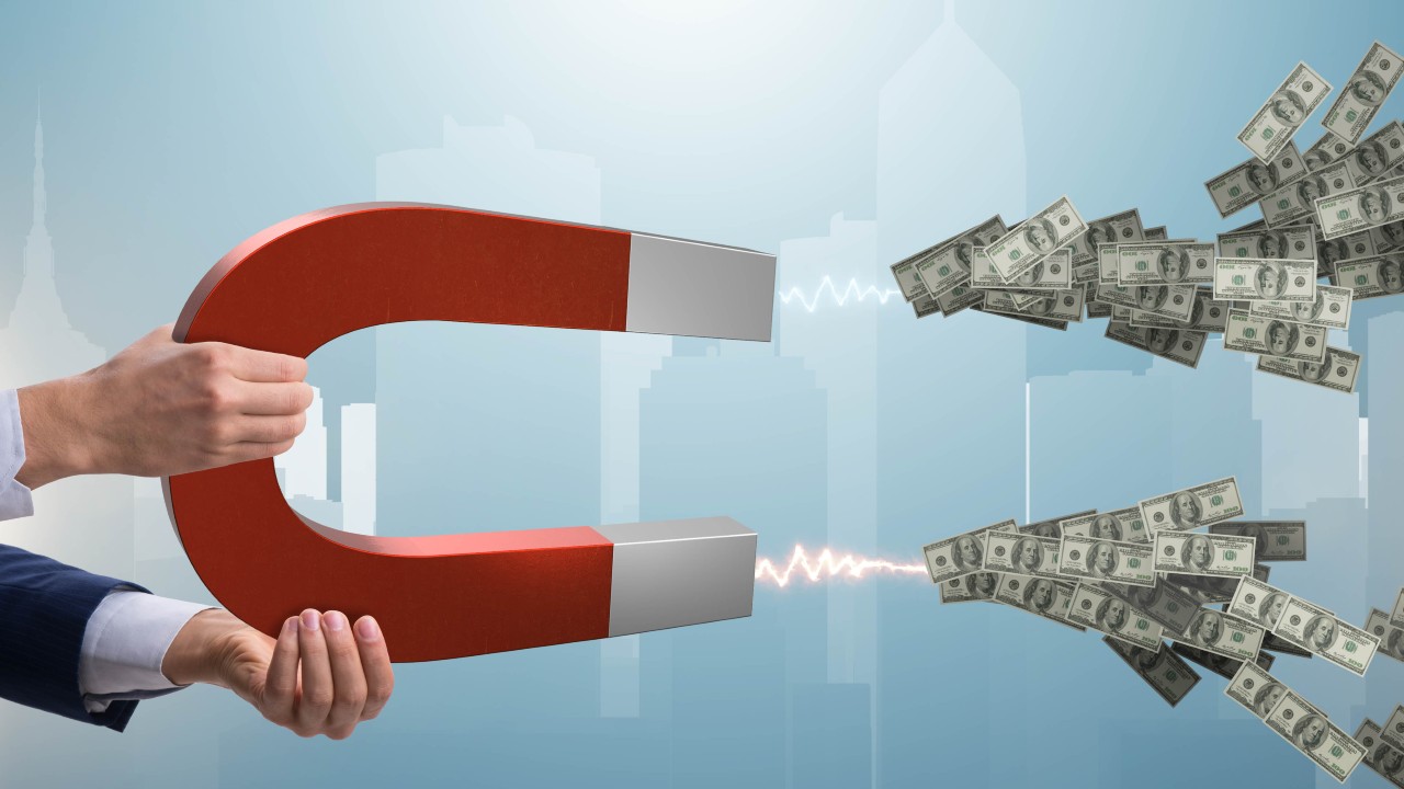 Una persona tiene in mano un grosso magnete rosso che attrae pile di banconote da un dollaro, simboleggiando l'attrazione della flat tax per la ricchezza finanziaria.