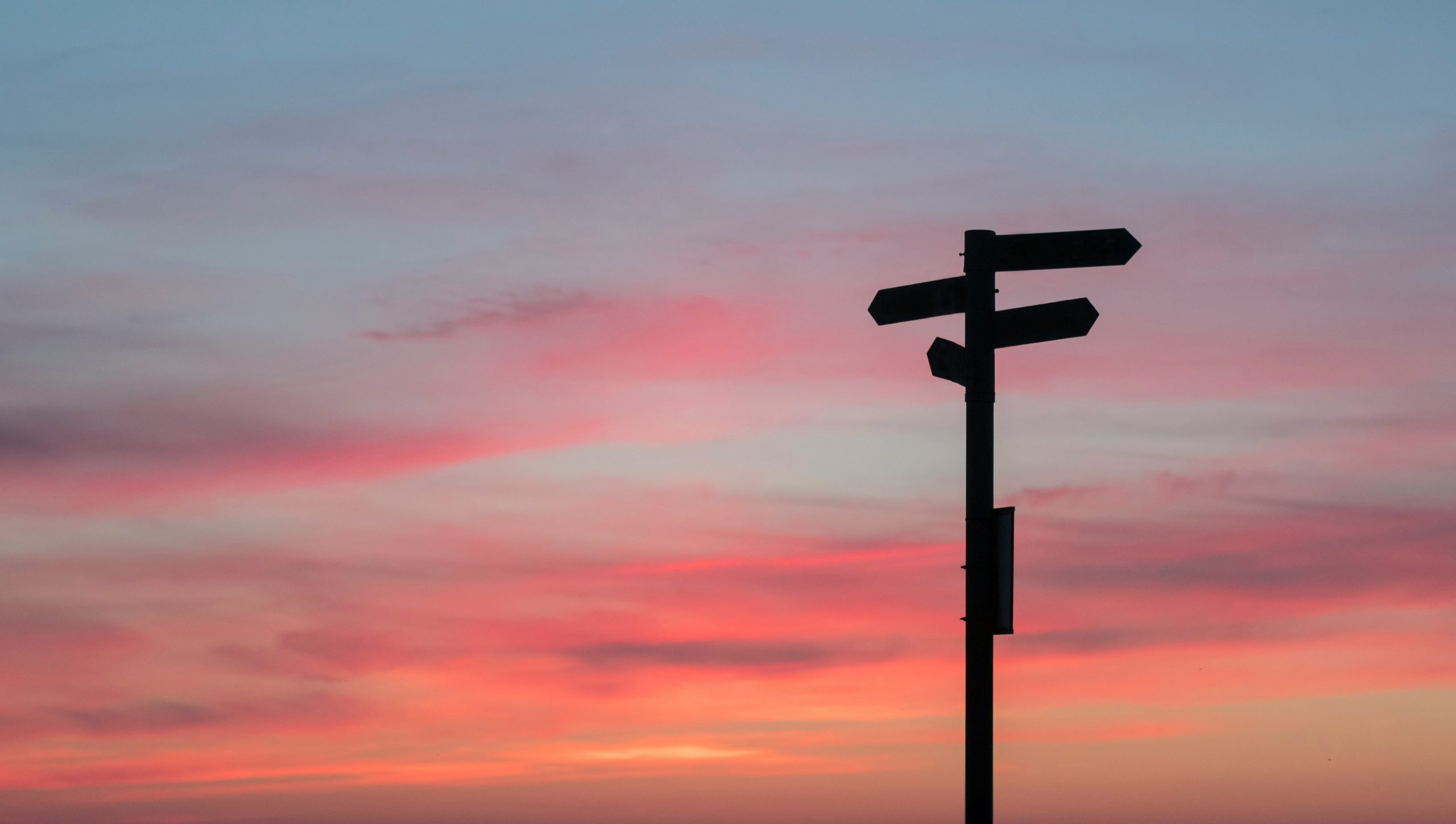La sagoma di un cartello stradale con più frecce direzionali si staglia contro un cielo colorato al tramonto, ricco di sfumature rosa, arancioni e viola.