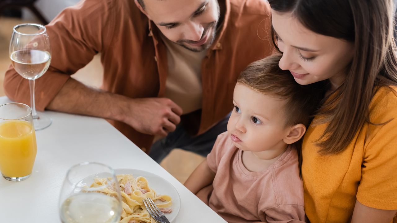 Una famiglia di tre persone è seduta a un tavolo da pranzo. Il padre e la madre sorridono mentre guardano il loro bambino, concentrato su un piatto di pasta. Bicchieri di succo e vino sono disposti accanto all'ultimo opuscolo "legge di bilancio", aggiungendo all'atmosfera calda e intima.