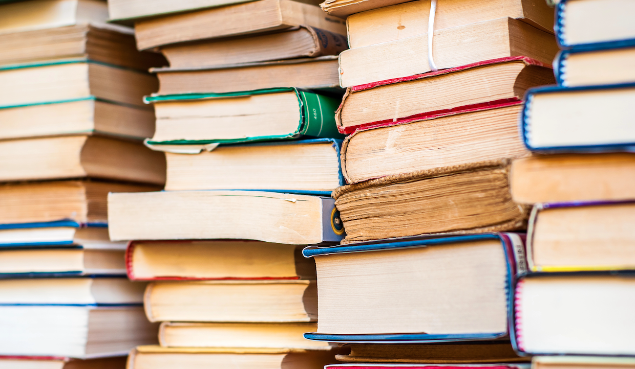 Pile di libri colorati ammucchiati uno vicino all'altro, con dorsi e pagine visibili. I libri hanno spessori diversi e mostrano segni di usura, che indicano un uso frequente. Lo sfondo è parzialmente sfocato, enfatizzando i libri in primo piano.