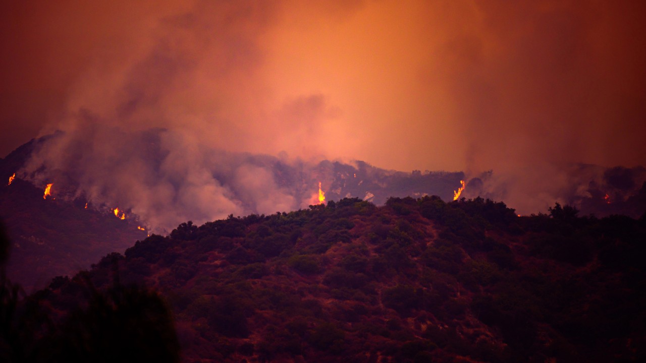 Un incendio boschivo divampa in una zona montuosa, proiettando un drammatico bagliore arancione nel cielo. Un denso fumo si solleva verso l'alto, con fiamme visibili lungo la cresta, in contrasto con la sagoma scura delle montagne.