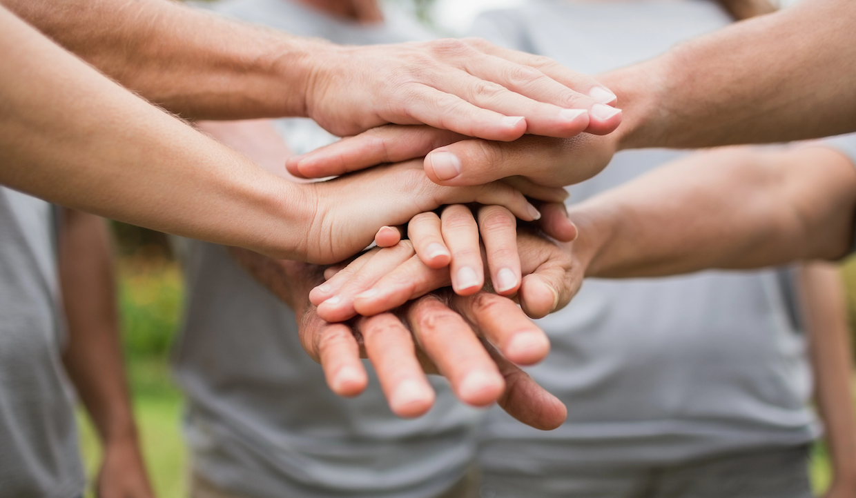 Un gruppo di persone in piedi insieme, ciascuna con una mano sopra l'altra in una pila, a simboleggiare unità e lavoro di squadra. Sono all'aperto e indossano abiti casual.