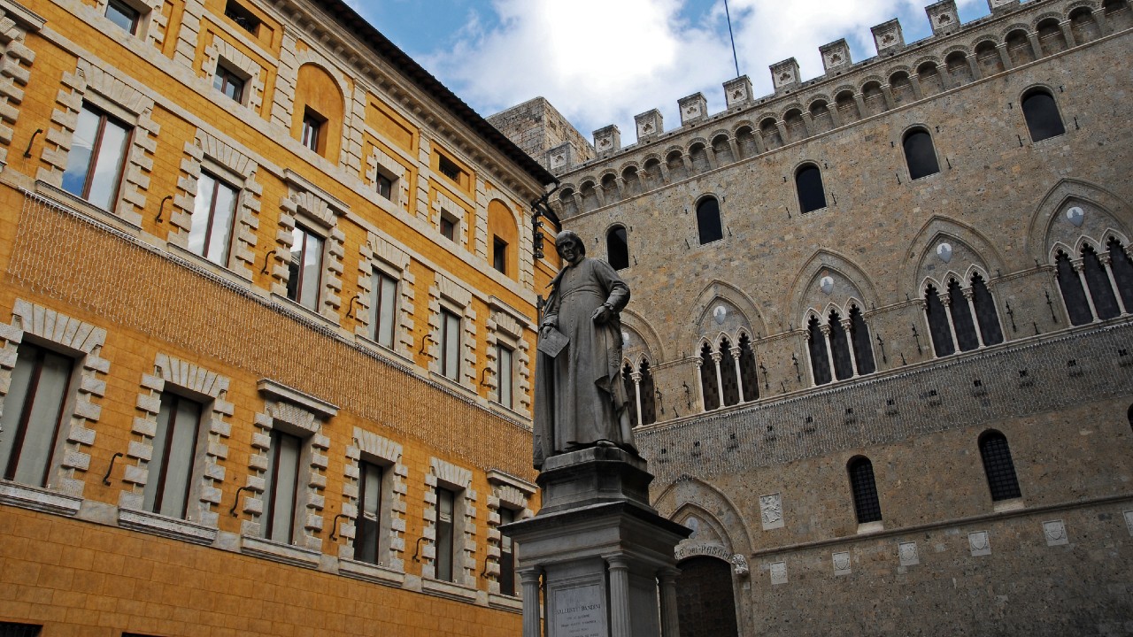 Una statua si erge in una piccola piazza, circondata da edifici storici con facciate ornate. Un edificio è giallo con accenti bianchi, mentre l'altro è grigio con archi e una merlatura. Il cielo è parzialmente nuvoloso.
