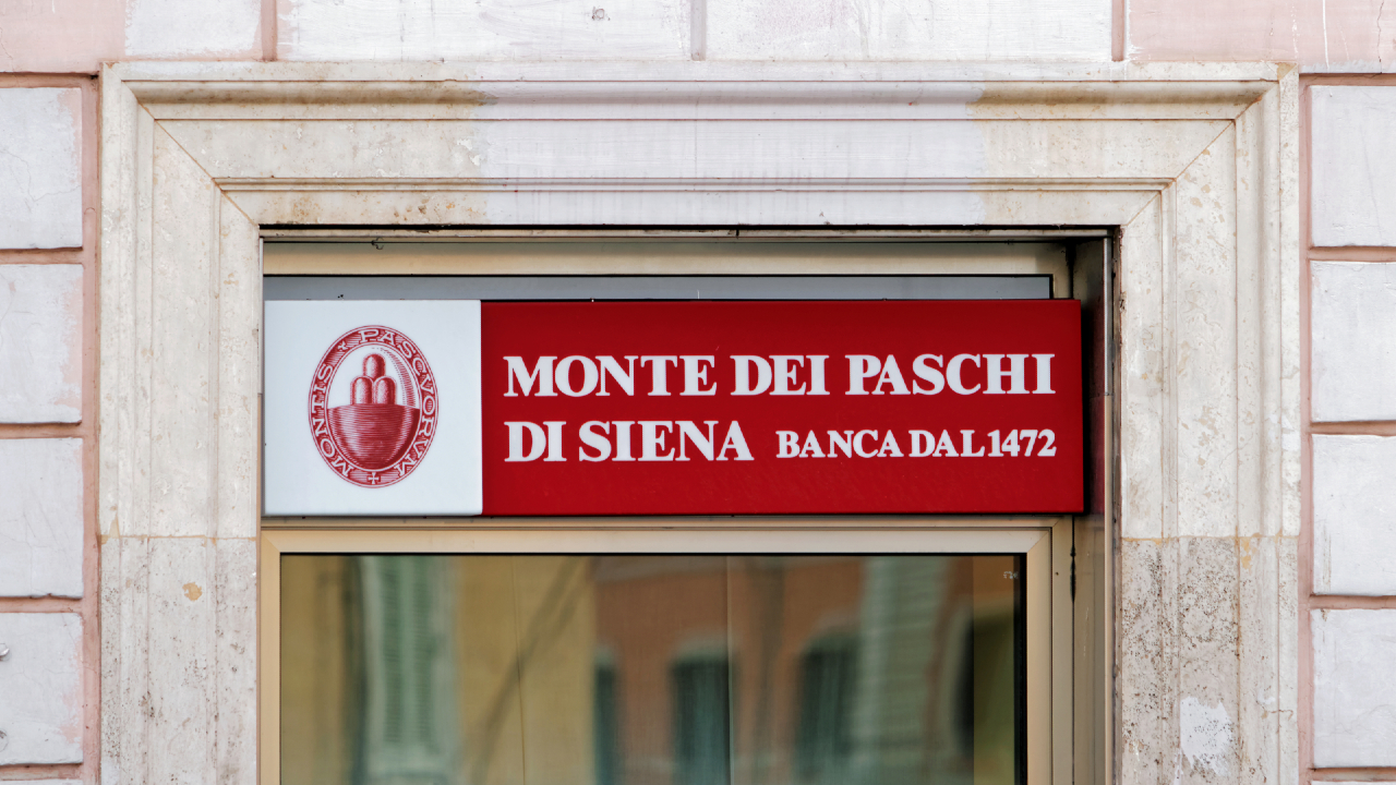 Insegna per Monte dei Paschi di Siena, che mostra il testo "Banca dal 1472" in bianco su sfondo rosso, con uno stemma sulla sinistra. L'insegna è sopra una porta a vetri incorniciata da un arco in pietra.