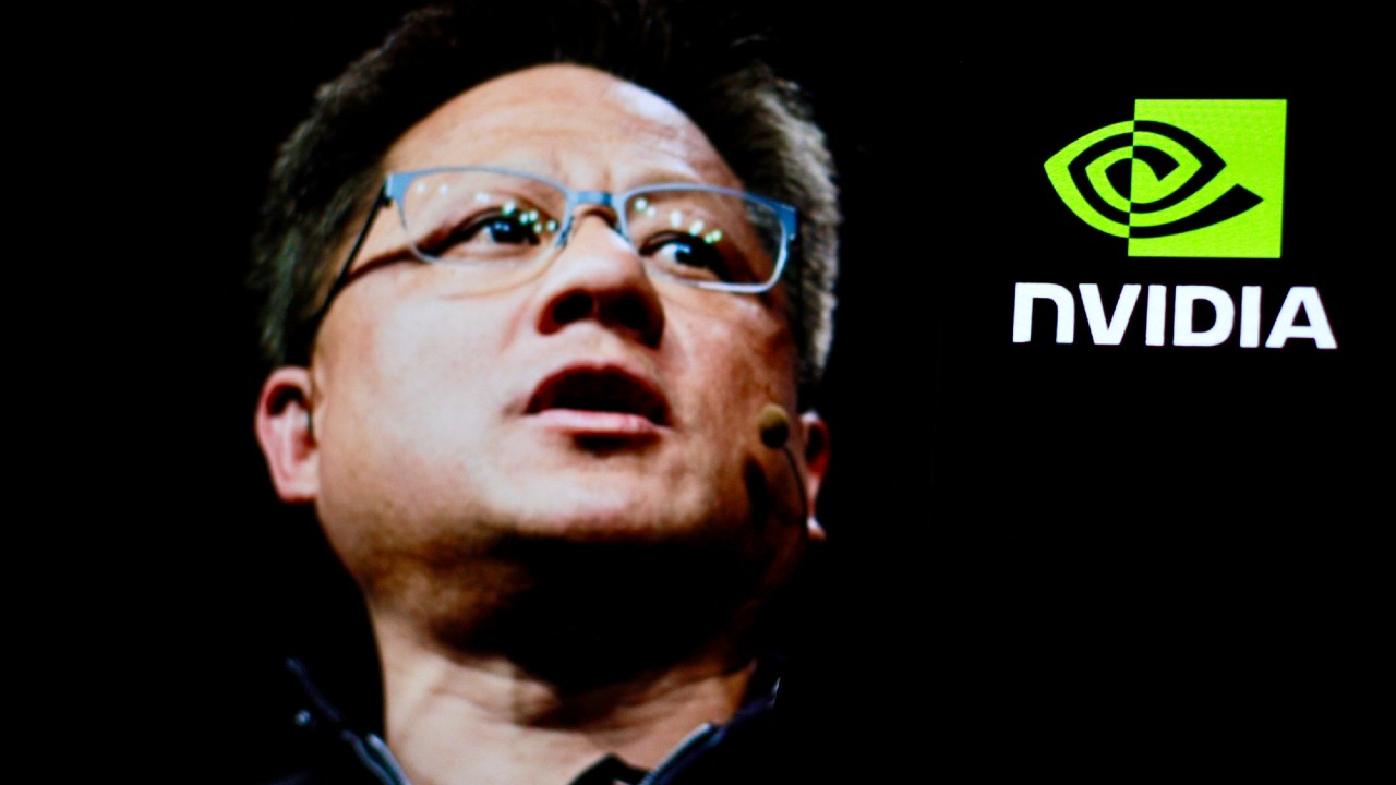 Uomo che parla sul palco con un microfono in cuffia, indossando occhiali. Il logo Nvidia appare su uno sfondo verde nell'angolo in alto a destra. L'illuminazione è fioca, evidenziando il suo volto.