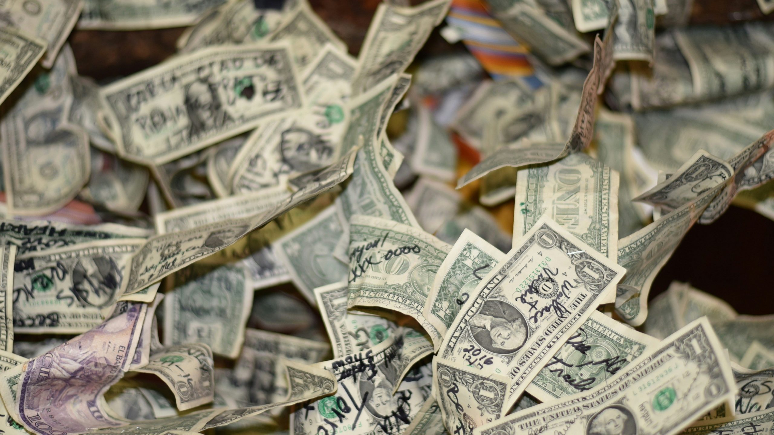 Un mucchio di banconote da un dollaro americane accartocciate, molte delle quali recano annotazioni e scarabocchi scritti a mano con vari inchiostri, sparse alla rinfusa.