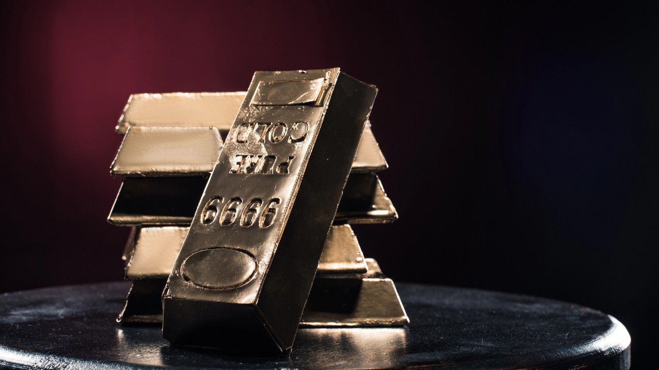 Una pila di lingotti d'oro è posta su una superficie scura su uno sfondo scarsamente illuminato. Un lingotto d'oro è posizionato in verticale di fronte, con numeri e lettere in rilievo sulla sua superficie. L'illuminazione mette in risalto la lucentezza metallica dei lingotti.