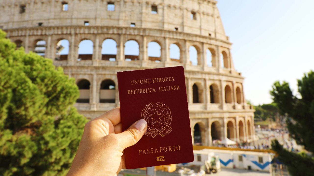 Una mano che tiene un passaporto italiano di fronte al Colosseo a Roma. L'antico anfiteatro, parzialmente visibile e immerso nella luce del sole, si erge maestoso sullo sfondo. Il verde incornicia l'angolo in basso a sinistra dell'immagine.
