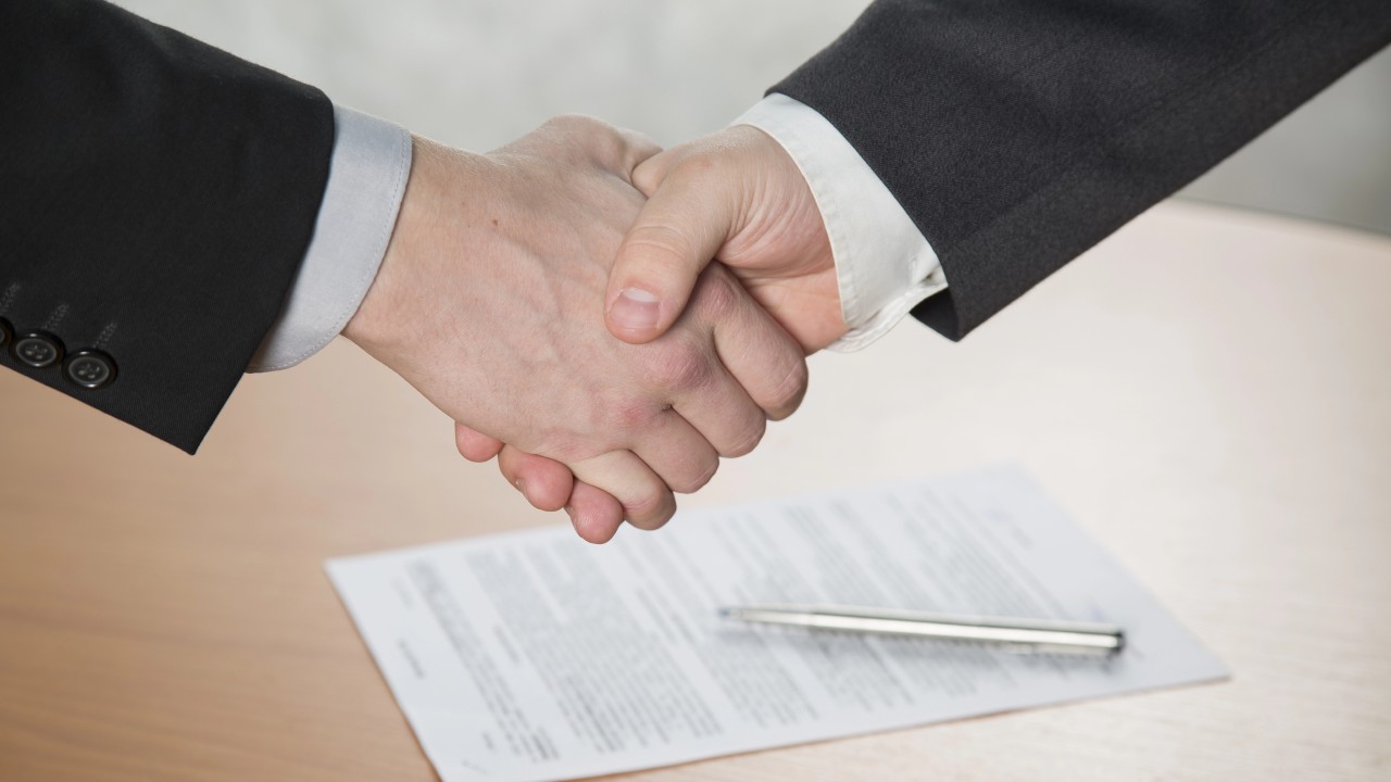 Due persone in abiti da lavoro si stringono la mano su un tavolo di legno con un documento e una penna sopra. L'immagine suggerisce un accordo o un affare commerciale in corso, come un patto di famiglia.