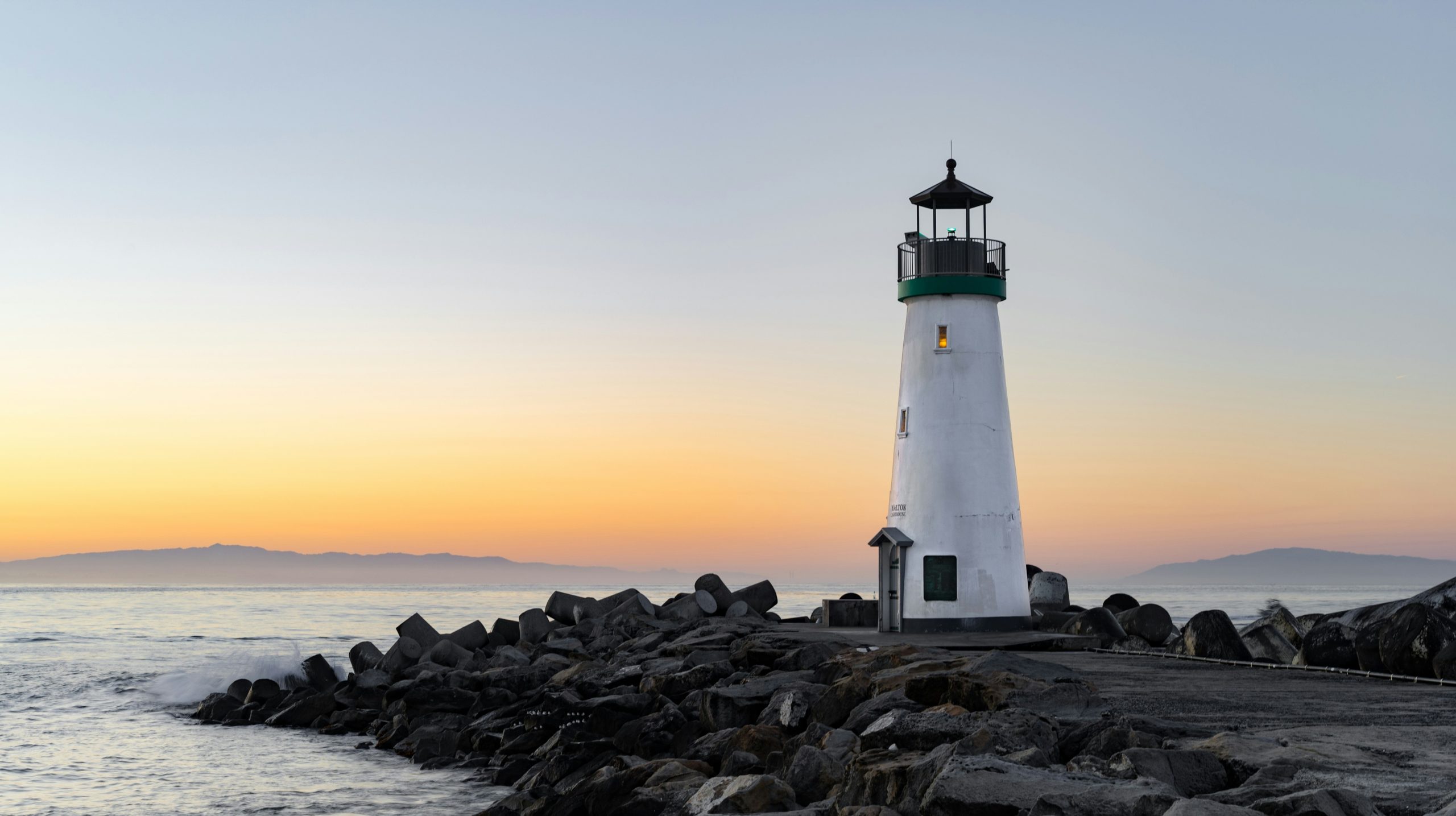 Un faro bianco con la cima verde si erge su una costa rocciosa durante il tramonto, come un guardiano per il portafoglio Carmignac. L'oceano calmo si estende fino all'orizzonte sotto un caldo cielo arancione e giallo, mentre le montagne lontane sussurrano debolmente storie per le generazioni future.