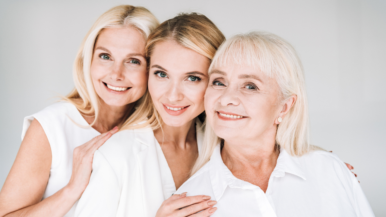 Tre donne di età diverse, sorridenti e vestite con camicette bianche, sono in piedi, vicine l'una all'altra, su uno sfondo chiaro, trasmettendo calore e un senso di connessione che ricorda la condivisione di storie in un'incantevole pensione.