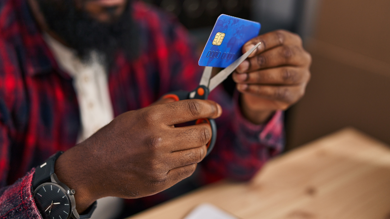 Una persona con una camicia a quadri rossa usa le forbici per tagliare una carta di credito "Conto" blu con un chip visibile. La foto si concentra sulle sue mani e sulla carta, con parte del suo viso e un tavolo di legno sullo sfondo.