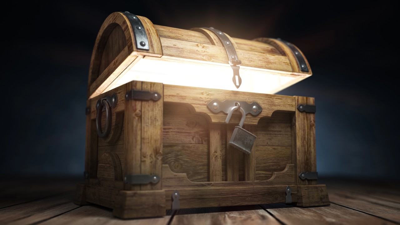 Uno scrigno del tesoro in legno leggermente aperto, che emette una luce brillante. Presenta cerniere in metallo e un lucchetto appeso sul davanti. Lo scrigno è posto su una superficie di legno con uno sfondo scuro, creando un'atmosfera misteriosa. L'immagine è per riprendere il tema della ricchezza nelle successioni e donazioni