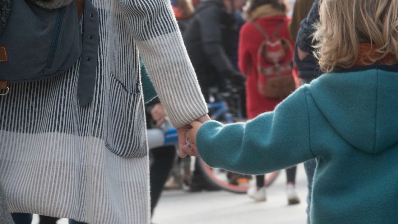 Un bambino e un adulto si tengono per mano in mezzo alla folla. Il bambino indossa un cappotto verde acqua, mentre l'adulto ne sfoggia uno a righe. Le persone sullo sfondo sono leggermente sfocate, con parte di una bicicletta visibile. L’idea è di collegarlo al tema della tutela dei minori nelle successioni.