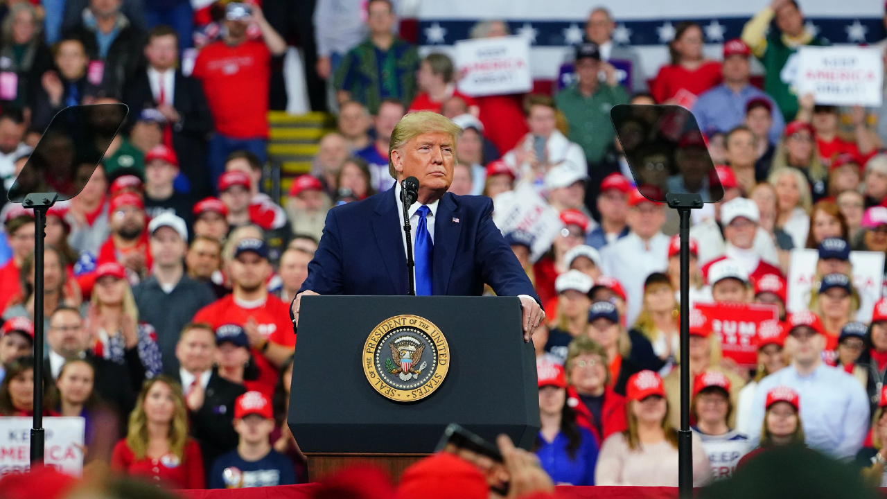 Una persona in abito scuro e cravatta blu, che ricorda lo stile di Trump, parla su un podio con un sigillo presidenziale. Lo sfondo rivela una folla numerosa e eterogenea di persone vestite di rosso, che agitano cartelli con entusiasmo.