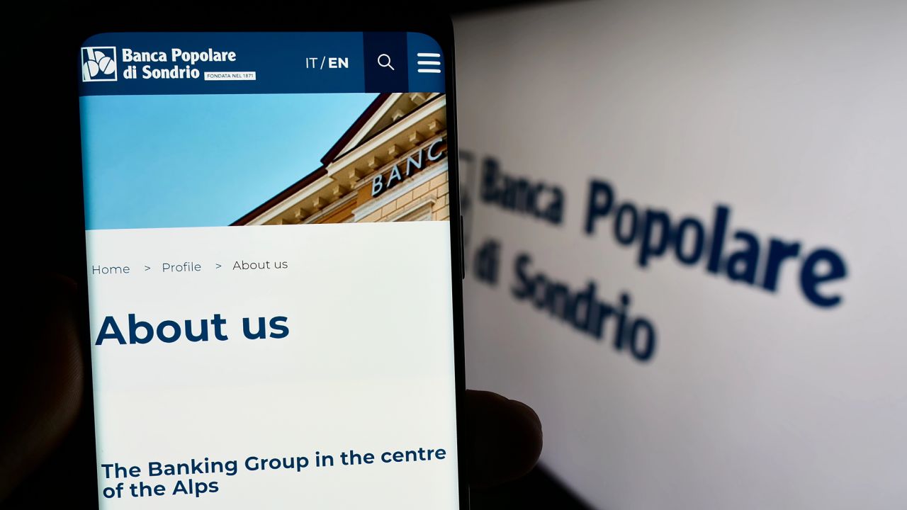 Uno smartphone visualizza la pagina "Chi siamo" del sito web della Banca Popolare di Sondrio, con in evidenza "Il Gruppo bancario al centro delle Alpi". Sullo sfondo di uno schermo sono visibili il logo e il nome della banca.