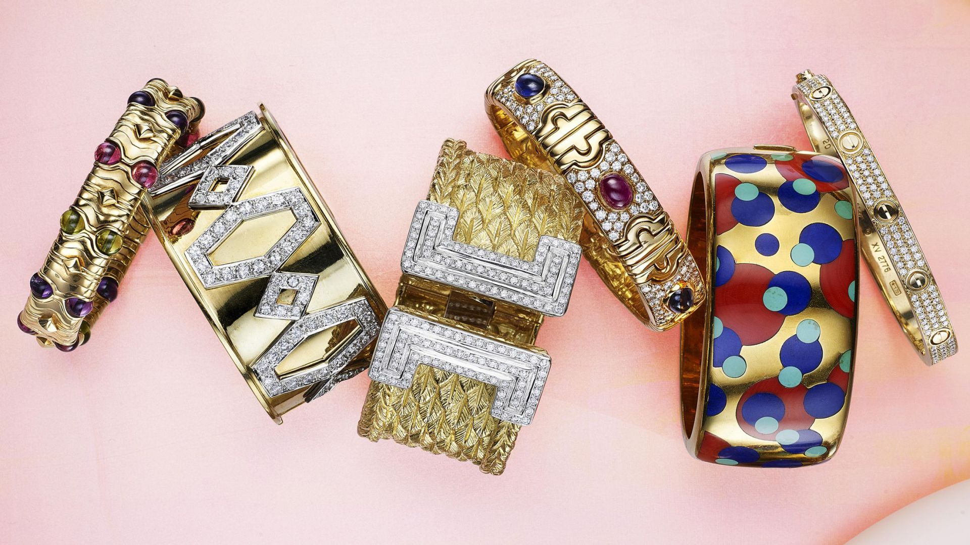 Una varietà di bracciali in oro e argento su uno sfondo rosa tenue. Presentano disegni intricati con pietre preziose, diamanti e motivi colorati. Ogni bracciale ha uno stile unico, dalle forme geometriche ai pois.