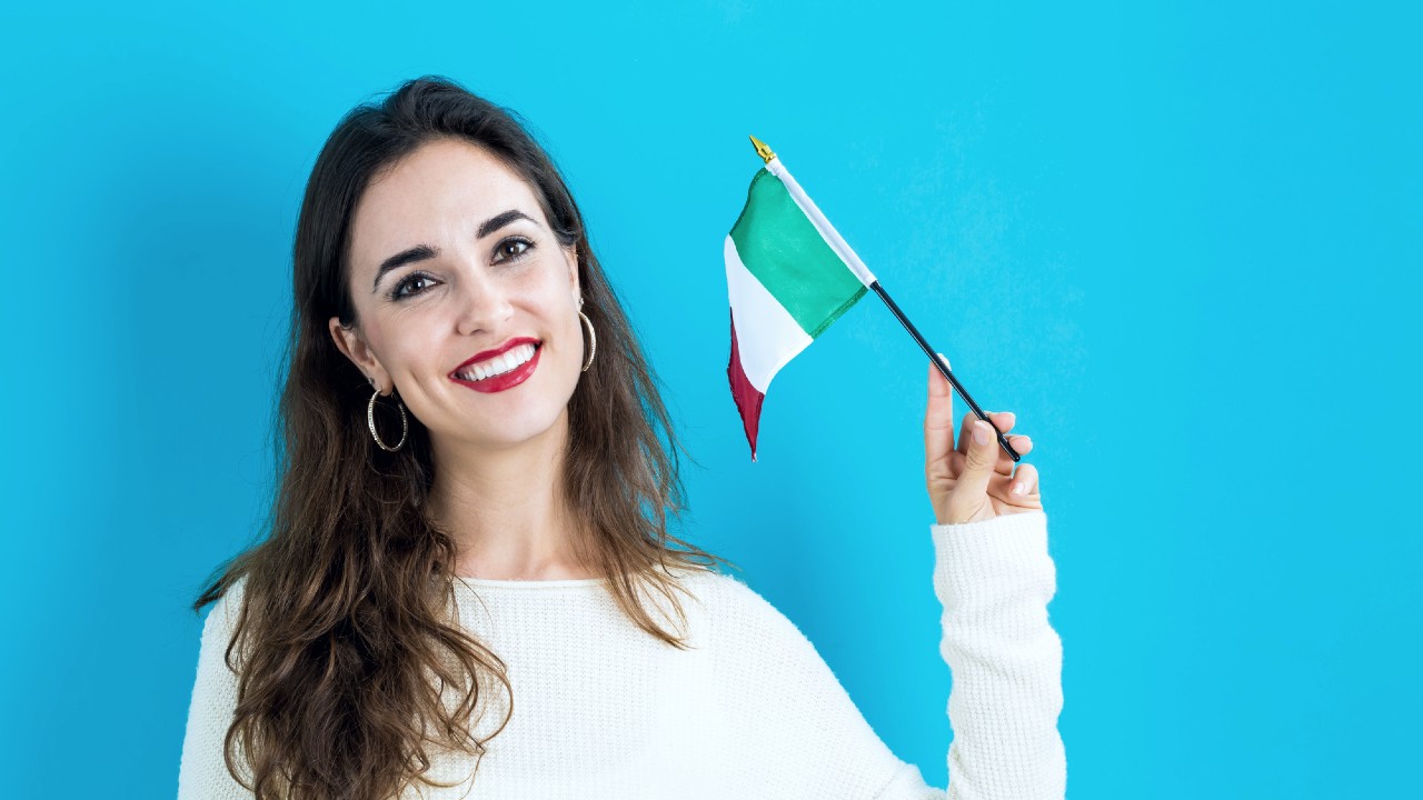 Una donna sorridente con i capelli lunghi tiene in mano una piccola bandiera italiana su uno sfondo blu brillante, indossa un maglione bianco e orecchini a cerchio.