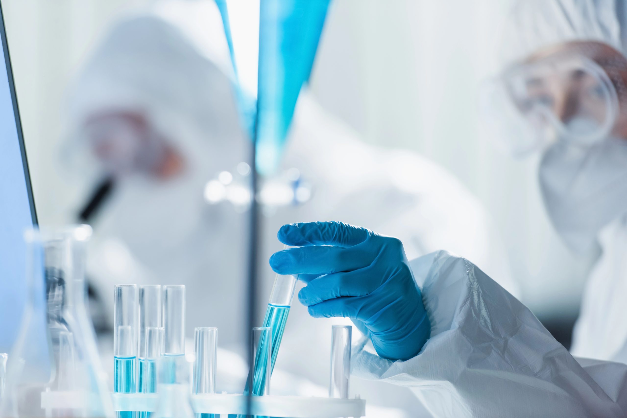 Gli scienziati in tute protettive lavorano meticolosamente in un laboratorio biotecnologico, concentrandosi su provette piene di liquido blu. Una persona regola una provetta mentre un'altra scruta attraverso un microscopio. L'ambiente è pulito e ben illuminato, sottolineando l'ambiente sterile essenziale per la ricerca avanzata.