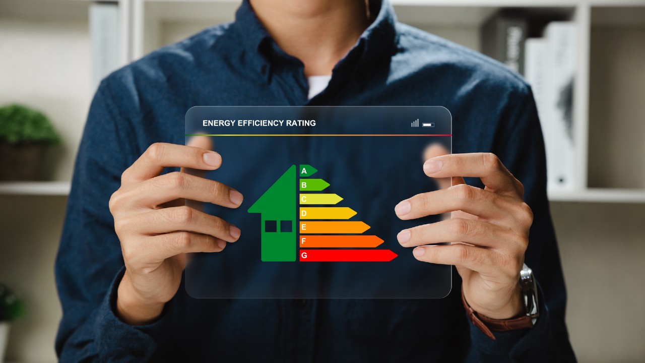 Una persona tiene in mano un tablet trasparente che mostra un grafico di valutazione dell'efficienza energetica a forma di casa con livelli che vanno da A (verde) a G (rosso). Sullo sfondo sono visibili scaffali con libri e una pianta.