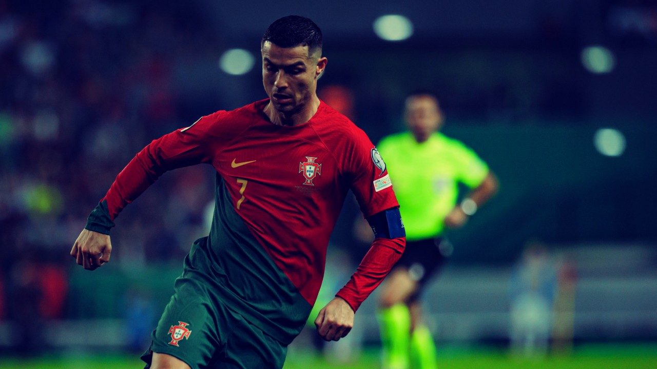 Un calciatore con la maglia rossa e verde del Portogallo con il numero 7 palleggia su un campo. Lo sfondo sfocato mostra un arbitro in uniforme luminosa e un pubblico in penombra.