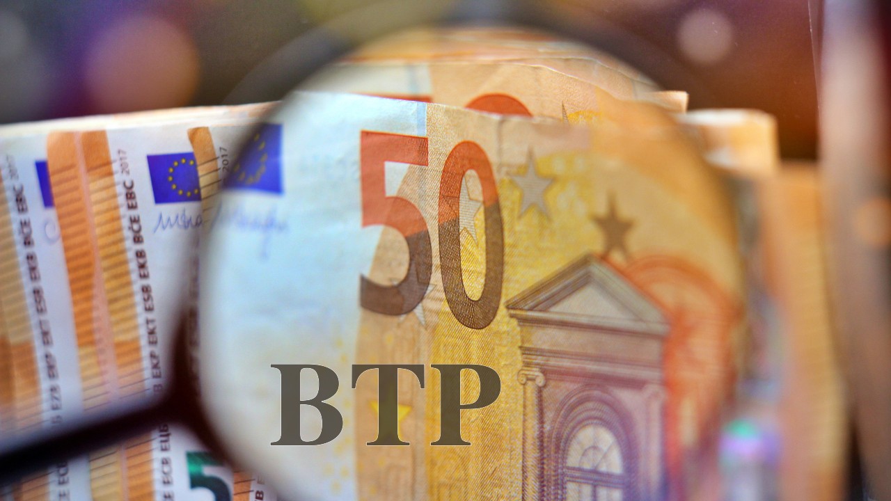 Una lente di ingrandimento si concentra su una pila di banconote da 50 euro, evidenziando le lettere "BTP". Le banconote sono parzialmente sfocate e sul bordo sinistro è visibile la bandiera dell'Unione Europea.
