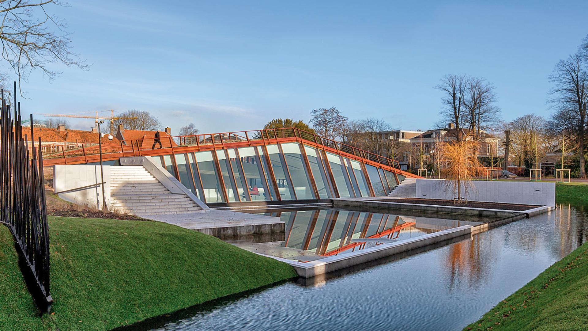 Un edificio moderno con una facciata triangolare in vetro parzialmente sommersa in un paesaggio erboso. Un canale d'acqua scorre lungo di esso, riflettendo la struttura. L'edificio ha una struttura color ruggine-arancione e il cielo è limpido con alcuni alberi sullo sfondo.