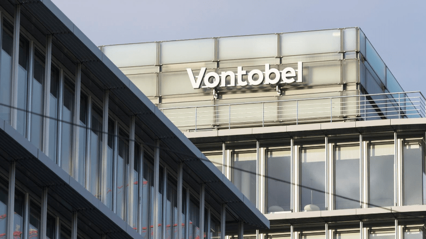Il moderno edificio in vetro mostra con orgoglio il logo "Vontobel", a testimonianza della prestigiosa filiale Vontobel Wealth Management Italia. È circondato da un'altra struttura in vetro riflettente, creando una nitida scena architettonica urbana sotto il cielo terso.