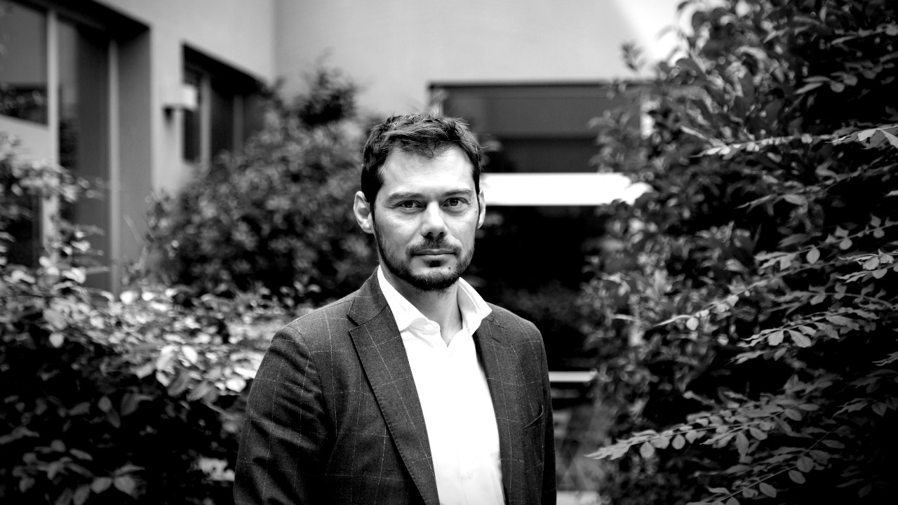 Un uomo in giacca e cravatta è in piedi all'aperto, circondato dal verde. L'immagine è in bianco e nero, il che le conferisce un tono classico e serio. Lo sfondo presenta cespugli e un edificio.