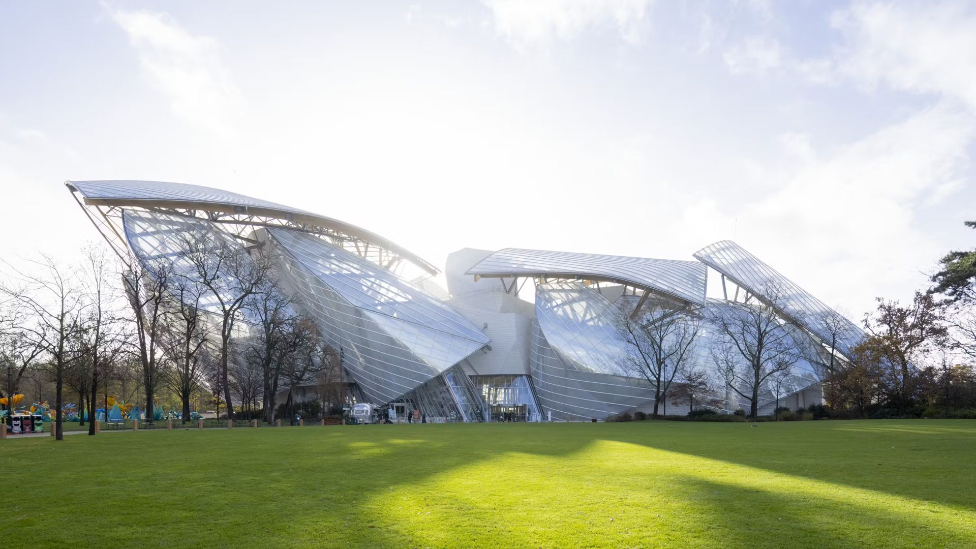 Edificio moderno con grandi pannelli di vetro angolari e strutture metalliche, situato in un parco con erba verde e alberi spogli. La luce del sole filtra attraverso le nuvole sopra, illuminando l'esclusivo design architettonico.
