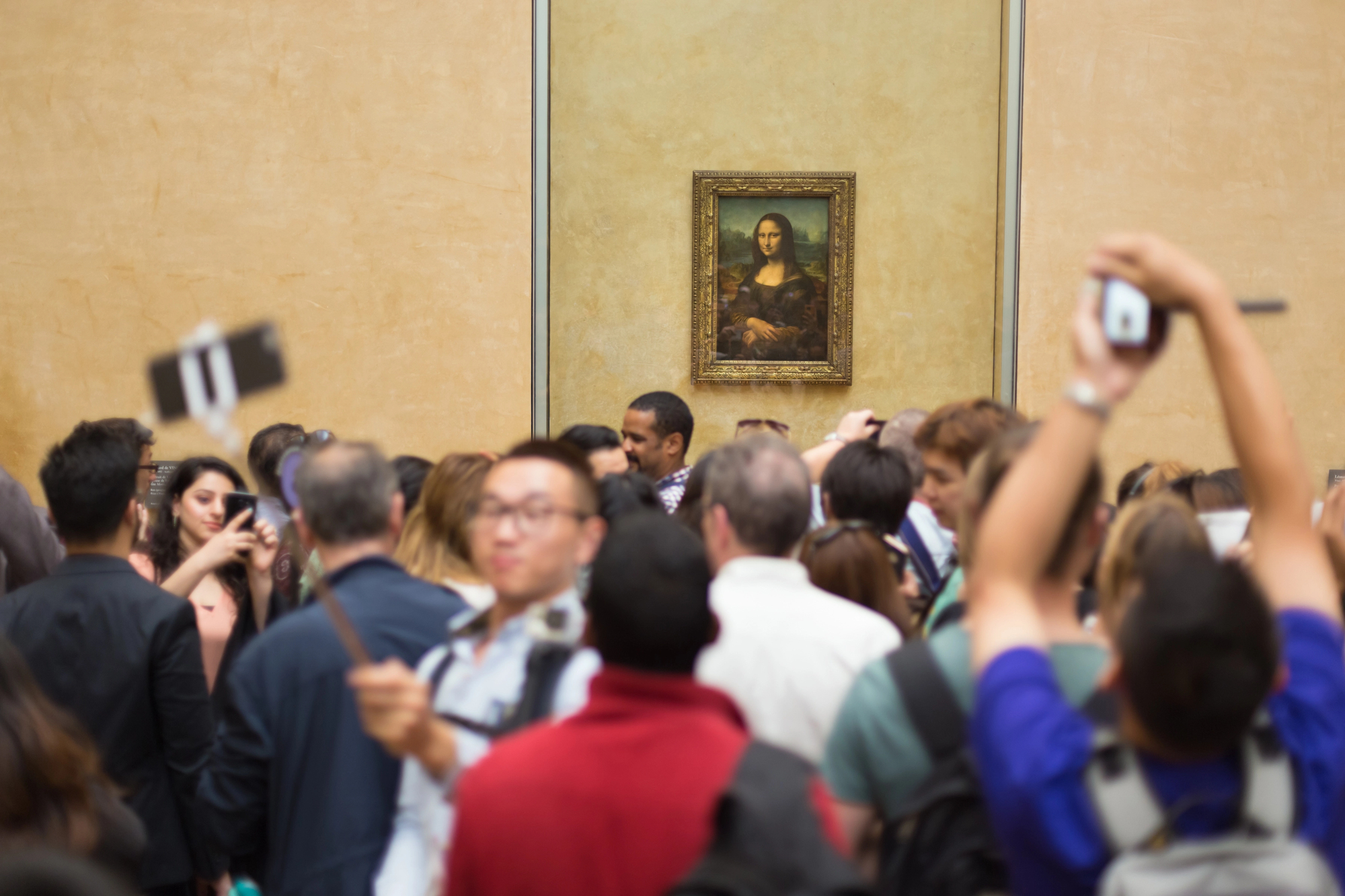 Una grande folla di persone si raduna attorno al dipinto della Monna Lisa in un museo. Molti tengono in mano telefoni e macchine fotografiche per scattare foto. La famosa opera d'arte è montata su una parete beige dietro una teca di vetro protettiva.