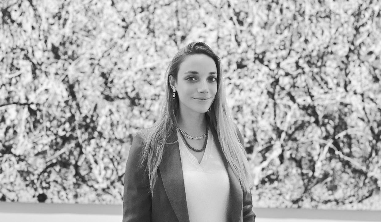 Marta Zava con i capelli lunghi e un tailleur è in piedi davanti a un dipinto astratto. L'opera d'arte dietro di lei appare intricata con un mix di motivi neri, bianchi e grigi. L'immagine è in bianco e nero.