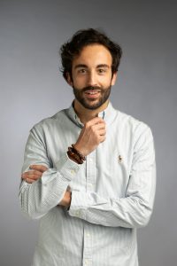 Un uomo con barba e capelli ricci è in piedi su uno sfondo grigio. Indossa una camicia abbottonata di colore chiaro e un braccialetto di perline, incrocia un braccio e si tocca il mento con l'altra mano.