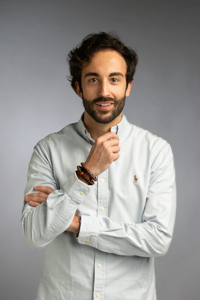 Un uomo con barba e capelli ricci è in piedi su uno sfondo grigio. Indossa una camicia abbottonata di colore chiaro e un braccialetto di perline, incrocia un braccio e si tocca il mento con l'altra mano.