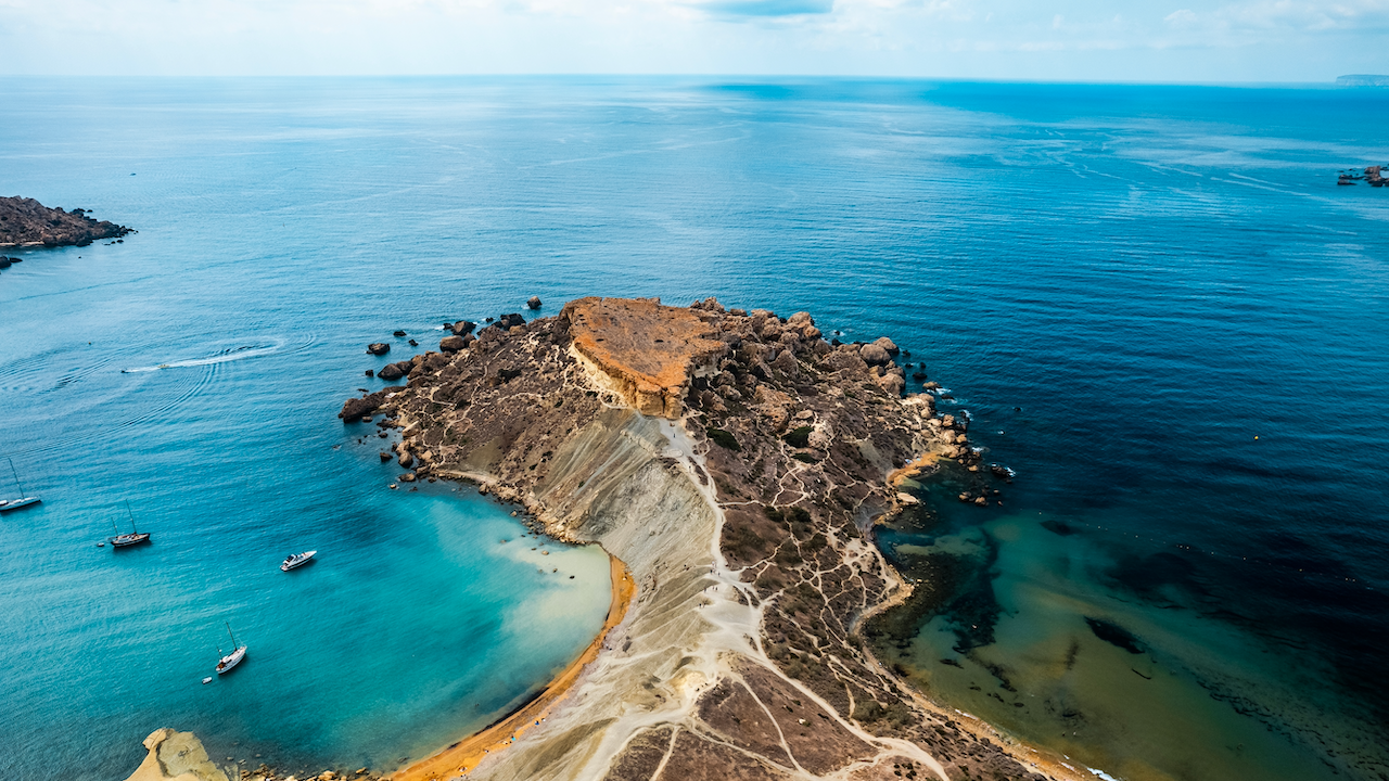 Vista aerea di una penisola rocciosa maltese con uno stretto sentiero sabbioso che si estende nell'oceano. Il mare circostante è un gradiente di blu e turchese. Diverse barche sono ancorate vicino alla costa. Il cielo è parzialmente nuvoloso con luce soffusa e diffusa.