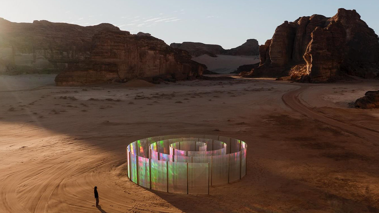 Una persona è in piedi vicino a un labirinto di vetro circolare e iridescente in un paesaggio desertico, circondato da scogliere rocciose. Il cielo è limpido e la luce del sole proietta lunghe ombre sulla sabbia.