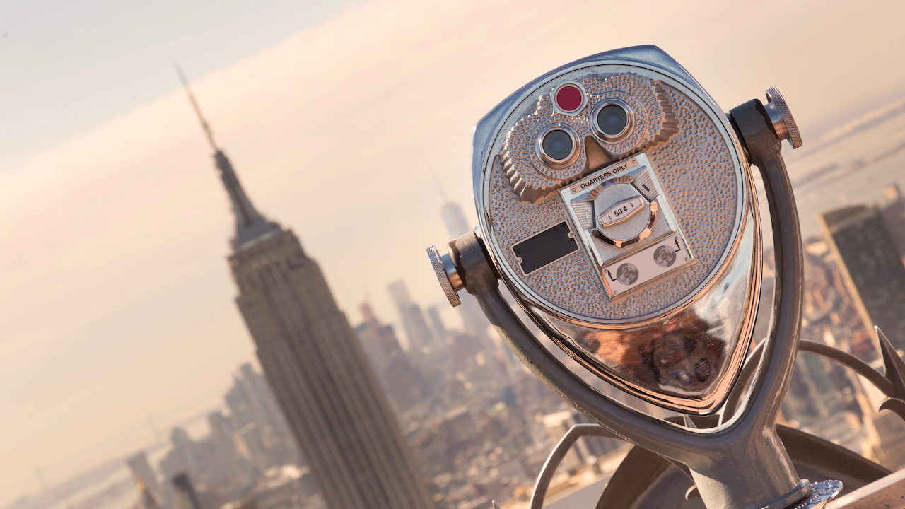 Perchè investire in obbligazioni high yield. Un visore binoculare a gettoni si affaccia su un paesaggio urbano sfocato con l'Empire State Building ben visibile. Il visore è metallico e strutturato, sullo sfondo di uno skyline nebbioso sotto una luce soffusa.